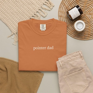 May include: An orange Comfort Colors t-shirt with the text "pointer dad" in white. The shirt is displayed with beige pants, a woven placemat, a candle, and a beige knitted throw.