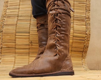 May include: Brown leather knee-high boots with a lace-up design. The boots have a pointed toe and are made of distressed leather. Braided leather detail wraps around the top of the boot. The boots are on a bamboo mat.