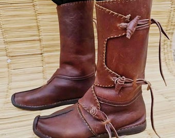 May include: A pair of brown leather boots with a unique design. The boots have a tall shaft, square toe, and decorative stitching. They feature leather ties and a rustic, handcrafted appearance, perfect for historical reenactments or costume wear.