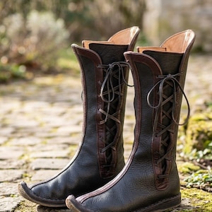May include: A pair of black leather boots with brown accents and laces. The boots have a tall, slender design with a curved toe and are set on a stone surface.