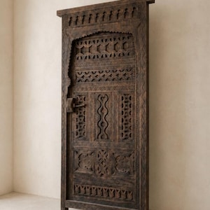 Pode incluir: Uma porta de madeira castanha escura, intrincadamente esculpida. A porta apresenta padrões detalhados, incluindo formas geométricas, arcos e motivos florais. A porta tem uma forma retangular com um topo decorativo e uma tranca.