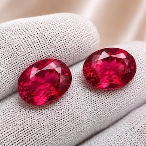 May include: Two oval-cut, deep red gemstones with faceted surfaces that reflect light. The stones are positioned on a textured, light gray fabric surface, displaying a vibrant crimson hue and a polished, transparent appearance throughout.