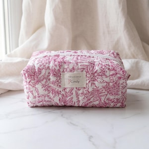 Pode incluir: Uma bolsa de cosméticos retangular acolchoada com fundo branco e estampa floral rosa. A bolsa tem um zíper branco e uma pequena etiqueta que diz "Fait avec amour" e "Cindy". A bolsa está em uma superfície de mármore branco.