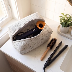 May include: A quilted beige toiletry bag with a small dark floral pattern, containing a dark blue and orange hair dryer. Beside the bag lies a round wooden hair brush with black bristles and a black hair straightener with a cord attached.
