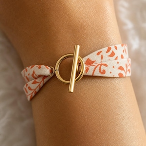 Peut inclure: Un bracelet composé d'une bande de tissu couleur crème avec un imprimé botanique orange. La bande est fixée par un fermoir à bascule en métal doré poli, composé d'un anneau circulaire et d'une barre droite, créant une fermeture simple.