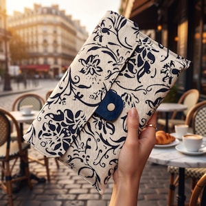 May include: A rectangular clutch bag made of beige fabric with a dark navy blue floral pattern. The bag features a fold-over flap secured by a small, dark navy blue tab with a single gold-toned metal snap button closure at the center front.