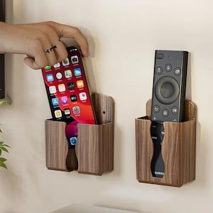 May include: Two wall-mounted storage holders with a brown wood grain finish. One holder contains a smartphone with a colorful app icon display, and the other holder contains a black remote control with various buttons and the Samsung brand name.