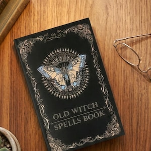 Op de afbeelding: Een zwart rechthoekig boek met de tekst OLD WITCH SPELLS BOOK in goud gedrukt. De kaft bevat een centrale illustratie van een vlinder met blauwe en beige vleugels, omringd door een zonnestraalpatroon en een gouden filigraanrand.
