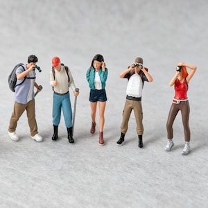 May include: A set of five miniature figurines depicting hikers in outdoor gear. The figures include individuals wearing backpacks, hats, and casual clothing, with accessories such as binoculars, cameras, and walking sticks, standing in a row.