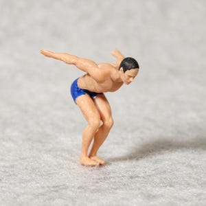 以下が含まれることがあります： 黒髪で青い水着を着用した、スタート姿勢をとる水泳選手のミニチュアフィギュア。両腕を外側に広げ、膝を曲げた姿勢で、水に飛び込む準備をしている動作を表現しています。