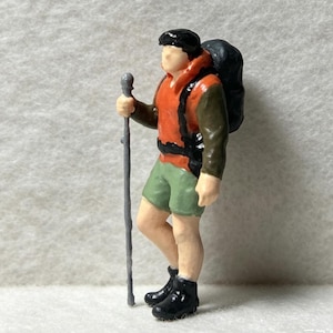 May include: A miniature figurine of a hiker with black hair, wearing an orange jacket, olive green long sleeves, and green shorts. The figure carries a large black backpack and holds a tall, thin gray walking stick. The hiker wears black boots.