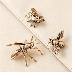 Puede incluir: Tres broches decorativos de bronce con forma de insectos. Los broches incluyen una abeja y una avispa, cada uno con alas y patas detalladas. Los broches están colocados sobre un fondo color crema.