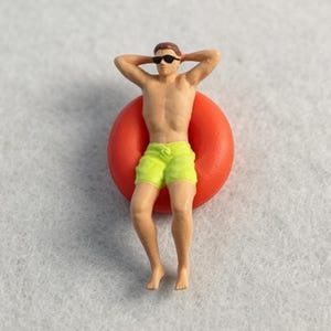 May include: A miniature figurine of a person with brown hair wearing black sunglasses and bright yellow swim trunks, resting with arms behind their head while floating inside a vibrant, solid orange circular inflatable pool ring float.
