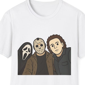 Può includere: Una t-shirt bianca a maniche corte con una grafica a cartoni animati di tre personaggi di film horror. La stampa mostra una figura con una maschera da fantasma nera, una figura con una maschera da hockey e giacca marrone, e una figura dai capelli scuri.