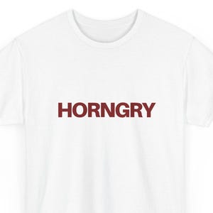 May include: A white short-sleeved t-shirt featuring the word HORNGRY printed in a bold, dark red sans-serif font across the center of the chest. The garment has a standard crew neckline and a straight hemline, presented on a plain white background.