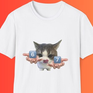 Peut inclure: Un t-shirt blanc à manches courtes avec une impression graphique représentant un chat aux mains humaines tendues vers l'extérieur. Le chat a une petite langue rose visible et est placé entre deux icônes carrées bleues affichant les chiffres 6 et 7.