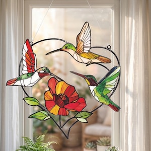 Peut inclure: Décoration de fenêtre en vitrail représentant des colibris dans un cadre en forme de cœur. Le design coloré présente trois colibris, une fleur rouge et jaune et des feuilles vertes. L'art de la fenêtre est suspendu par un fil fin.