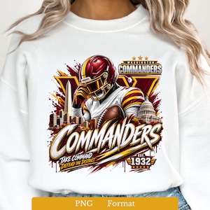 May include: A white sweatshirt featuring a graphic of a football player in a burgundy and gold helmet and jersey, holding a football. The design includes the text Washington Commanders, Take Command, Defend the District, and Est. 1932.