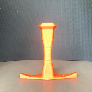May include: A bright orange, T-shaped stand featuring a vertical column with a textured spiral pattern on the front and a flat, horizontal base. The object has a smooth, matte finish and a symmetrical design with a wider top section.