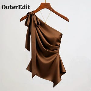 May include: A dark brown one-shoulder top with a satin-like finish, featuring a draped bodice and a long tie detail on the shoulder. The garment is displayed on a wooden hanger with a gold-tone necklace. The text OuterEdit is visible.