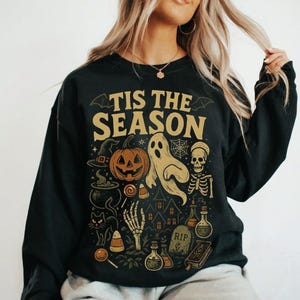 Puede incluir: Una sudadera negra de manga larga con el texto 'TIS THE SEASON' en dorado. El gráfico incluye una calabaza, un fantasma, un esqueleto, un sombrero de bruja, un caldero, un gato negro, una casa encantada, pociones, una lápida y maíz dulce.