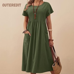 May include: An olive green midi dress featuring short sleeves, a round neckline, and vertical pleats across the bodice. The dress includes two side pockets and is accessorized with a long beaded necklace and a woven straw handbag.