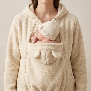 May include: A cream-coloured fleece hoodie featuring a central kangaroo-style pouch designed for carrying an infant. The pouch includes small decorative ears, and the infant is wearing a matching cream-coloured knit beanie with a small bow detail.