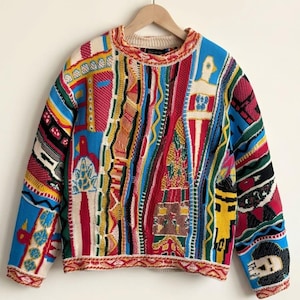 Puede incluir: Un suéter de manga larga y cuello redondo con un complejo patrón de punto multicolor. El diseño incorpora bloques vibrantes de rojo, azul, amarillo, verde y crema, con varias formas geométricas, figuras abstractas y rayas verticales texturizadas.