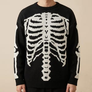 May include: A black long-sleeve knit sweater featuring a white graphic print of a human rib cage and spine on the front. The design extends to the sleeves, displaying white bone structures. The sweater has a ribbed crew neckline and cuffs.