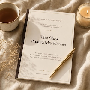 May include: A cream-colored planner titled The Slow Productivity Planner by Quietly Clear Studio, featuring dark brown text, a gold pen resting on the cover, a ceramic mug of tea, and a lit candle in a glass jar on a textured fabric surface.