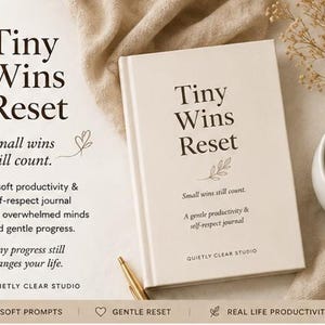 Może przedstawiać: Kremowy dziennik zatytułowany "Tiny Wins Reset" z tekstem "Small wins still count". Widoczny jest również złoty długopis i biały kubek z brązowym płynem. Dziennik przeznaczony jest do produktywności i szacunku do siebie.