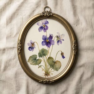 May include: An oval, antique gold-colored frame with a watercolor painting of purple violets. The painting features detailed leaves and flowers, set against a cream-colored background. The frame has ornate detailing and a small loop at the top for hanging.