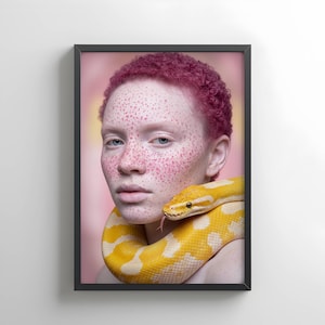 Puede incluir: Retrato enmarcado de una persona con cabello rosado y pecas, con una serpiente amarilla y blanca enrollada alrededor del cuello. Se ve la lengua de la serpiente. El fondo es rosa suave.