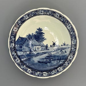 May include: A round ceramic bowl with a scalloped edge featuring a blue and white transferware pattern. The center depicts a rural landscape with a cottage, a river, and windmills. The rim is decorated with a repeating floral and scroll motif.