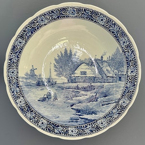 May include: A round ceramic plate with a scalloped edge featuring a blue and white transferware scene of a winter landscape with a thatched cottage, a windmill, a boat on a frozen canal, and two figures. The border has a floral scroll pattern.