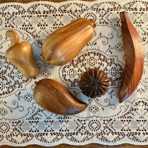 May include: A collection of wooden fruit and vegetable sculptures, including a banana, a pepper, a mango, a pumpkin, and a gourd. The sculptures are various shades of brown and sit on a white lace tablecloth.