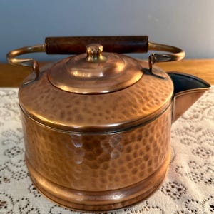 May include: A copper tea kettle with a hammered texture. The kettle has a dark wooden handle and a brass handle. The spout is on the right side of the kettle. The lid has a small knob on top.