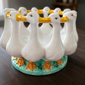 Peut inclure: Un bol en céramique présentant une disposition circulaire de canards blancs aux becs jaunes. Les canards sont reliés au niveau du cou, formant le bord du bol. La base est peinte avec de l'herbe verte et des pattes palmées orange.