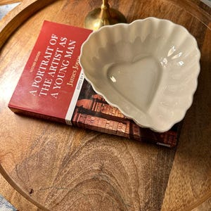 Può includere: Un piatto in ceramica a forma di cuore, bianco sporco, con un bordo smerlato, poggia su un libro intitolato "A Portrait of the Artist as a Young Man". Il libro è rosso con testo bianco. Gli oggetti sono su un vassoio di legno.