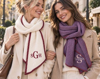 May include: Two ribbed knit scarves, one cream with maroon trim and the monogram SGH, the other purple with maroon trim and the name Sophia embroidered in white. Both scarves are worn over beige coats with matching sweaters.