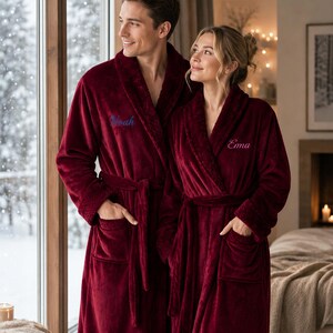 May include: Two plush, deep burgundy bathrobes featuring long sleeves, shawl collars, and tie-waist belts. One robe is embroidered with the name Noah in blue script, and the other is embroidered with the name Ema in pink script on the chest.