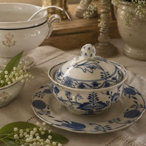 Op de afbeelding: Een witte porseleinen suikerpot met bijpassend deksel en schoteltje, voorzien van een blauw uienpatroon. De pot heeft een ronde vorm, een deksel met knop en een geschulpte rand aan het schoteltje. Het ontwerp bevat gedetailleerde blauwe bloemmotieven.