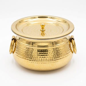 May include: A gold-colored, hammered-finish serving bowl with a matching lid. The bowl has two ring handles and a small knob on the lid. The bowl is round and appears to be made of metal.