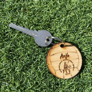 Könnte beinhalten: Ein Holzschlüsselanhänger mit einem Fußball-Design und dem eingravierten Namen "Aoife". Ein silberner Schlüssel liegt neben dem Schlüsselanhänger auf grünem Gras. Der Schlüsselanhänger hat auch eine Grafik und Text.