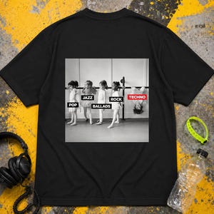 Può includere: Una t-shirt nera a maniche corte con una grafica centrale in bianco e nero di quattro bambini in abbigliamento da danza classica alla sbarra. La grafica include le etichette POP, JAZZ, BALLADS, ROCK e TECHNO sopra o vicino a ogni bambino.