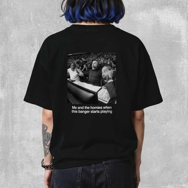 May include: Black t-shirt featuring a black and white image of people reacting with excitement. The text "Me and the homies when this banger starts playing" is below the image. The shirt is a casual, unisex style.