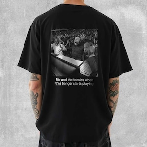 May include: Black t-shirt featuring a black and white image of three men reacting with excitement. The image is a meme with the text "Me and the homies when this banger starts playing". The shirt is a casual style.