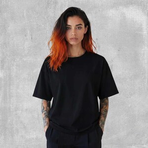 May include: A black short-sleeved t-shirt. The model is wearing a black t-shirt and black pants. The model has long hair with orange and black highlights. The background is a gray wall.