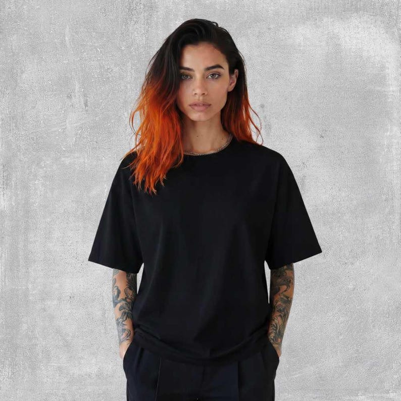 May include: A black short-sleeved t-shirt. The model is wearing a black t-shirt and black pants. The model has long hair with orange and black highlights. The background is a gray wall.