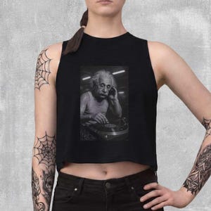 May include: Black crop top featuring a black and white image of Albert Einstein as a DJ. The image shows Einstein wearing a white tank top, with his hand on his ear and the other on a record player. The crop top is sleeveless.